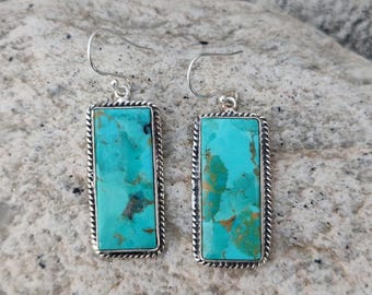 May include: A pair of turquoise earrings with a rectangular turquoise stone set in a silver frame. The frame has a twisted rope design around the edges. The earrings have a hook closure.