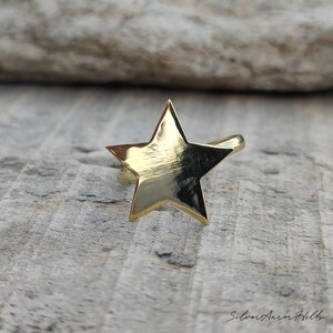 May include: A gold-toned ring featuring a prominent five-pointed star design on the front. The star has a smooth, reflective surface that catches the light. The band of the ring is visible extending from behind the star shape.