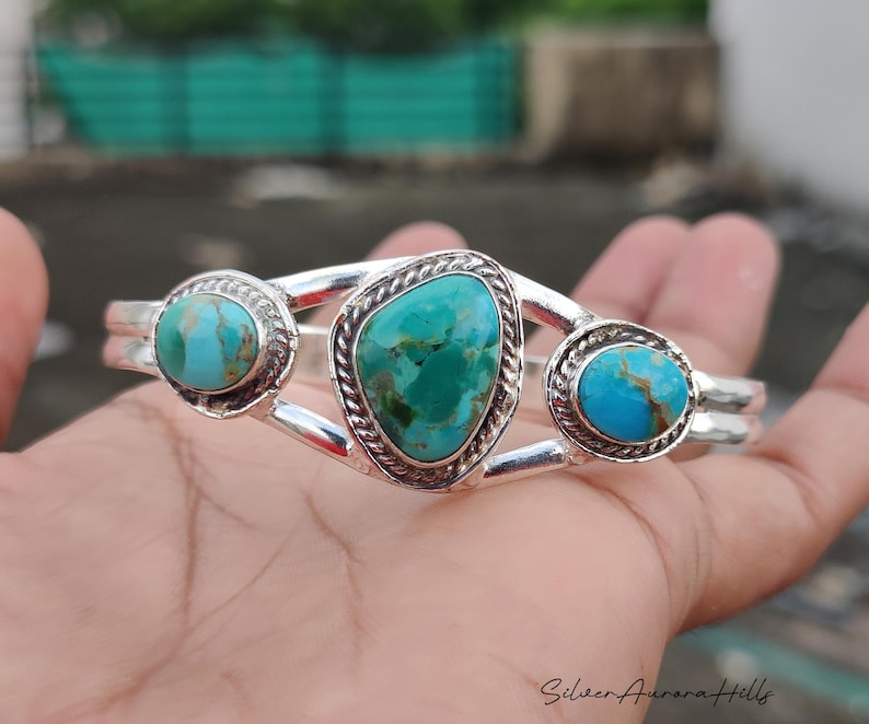 Op de afbeelding: Een zilveren armband met drie turkooizen stenen. De armband heeft een dubbele band met een grote, ovale turkooizen steen in het midden en twee kleinere, ronde stenen aan weerszijden. De stenen zijn gezet in decoratieve bezels.
