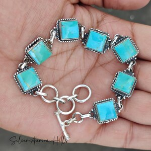 May include: A silver bracelet featuring square turquoise stones. Each stone is set in a square frame with a textured border. The bracelet has a toggle clasp and is displayed on a hand.