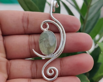 May include: A silver pendant featuring a polished, oval-shaped light green gemstone set within a curved, swirling metal frame. The design includes a prominent loop at the top for a chain and a decorative spiral at the bottom, crafted from polished silver.