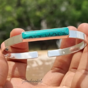 Könnte beinhalten: Ein silbernes Armband mit einem rechteckigen türkisfarbenen Stein in der Mitte. Das Armband hat eine polierte Oberfläche und ist unten offen. Der Text "SilverAuroraHills" befindet sich unten.
