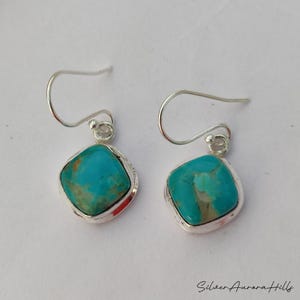 May include: A pair of turquoise earrings with a square, cushion-cut stone set in a silver frame. The earrings have a simple hook design. The turquoise stones have a mix of blue and green hues.