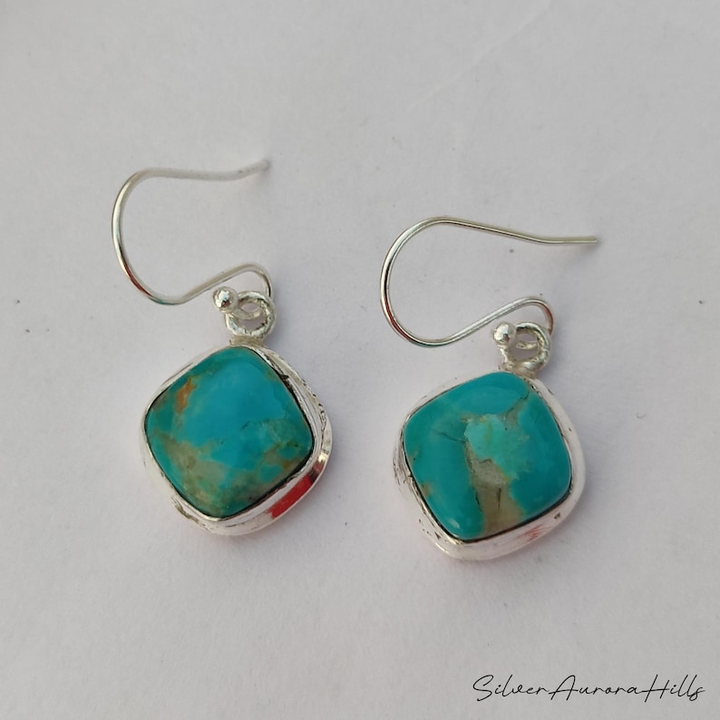 May include: A pair of turquoise earrings with a square, cushion-cut stone set in a silver frame. The earrings have a simple hook design. The turquoise stones have a mix of blue and green hues.