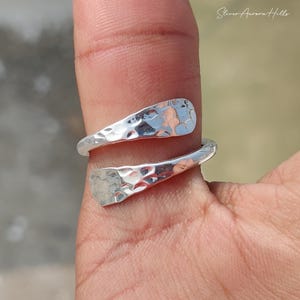 May include: A silver ring with a hammered texture, wrapped around a finger. The ring has a unique, organic shape and a bright, reflective surface. The ring is a piece of jewelry.