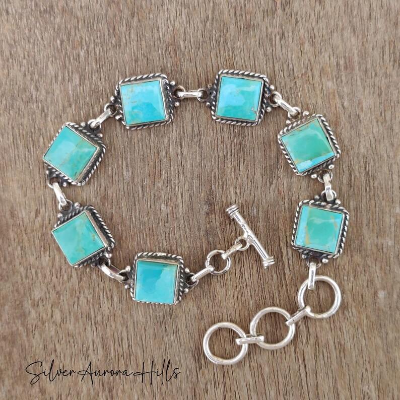 May include: A silver bracelet featuring square turquoise stones set in a decorative frame. The bracelet has a toggle clasp and is displayed on a wooden surface. The text "Silver Aurora Hills" is at the bottom.