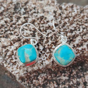 May include: A pair of turquoise and silver earrings. Each earring features a square, light blue turquoise stone set in a silver bezel. The earrings have a simple hook design. The earrings are displayed on a textured, brown surface.