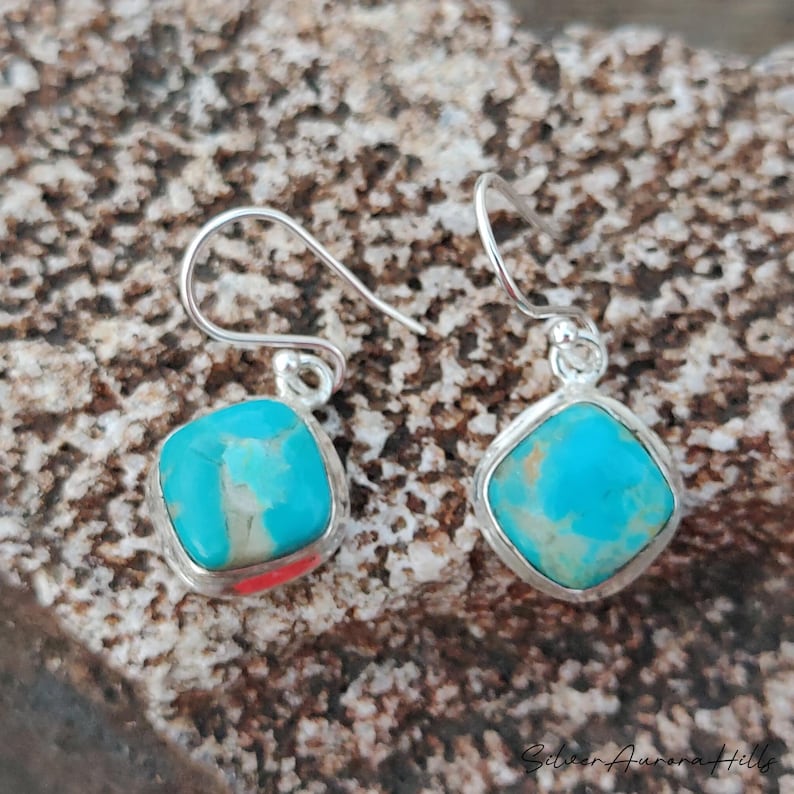 May include: A pair of turquoise and silver earrings. Each earring features a square, light blue turquoise stone set in a silver bezel. The earrings have a simple hook design. The earrings are displayed on a textured, brown surface.