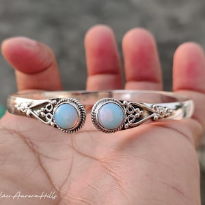 May include: A silver-toned cuff bracelet with two round, light blue opal stones. The bracelet features intricate filigree detailing and is open at the front. The text "Silver Aurora Hills" is at the bottom.