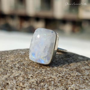 May include: A silver ring featuring a large, rectangular moonstone. The moonstone has a milky white base with iridescent blue and yellow flashes. The ring is set on a textured, neutral-toned surface.