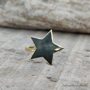 May include: A gold-toned ring featuring a prominent five-pointed star design on the front. The star has a smooth, reflective surface with visible fine brush-like textures. The band is thin and circular, connecting to the back of the star shape.