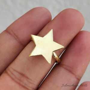 May include: A gold-toned ring featuring a prominent five-pointed star design on the front. The star has a smooth, flat surface and is attached to a simple, thin circular band that encircles the finger. The metal has a polished, reflective finish.