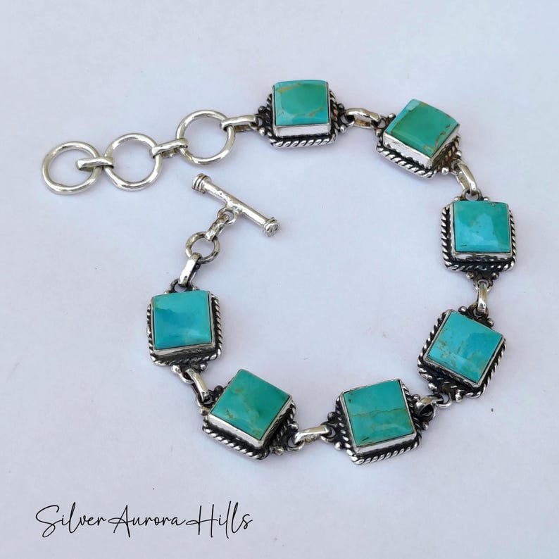 May include: A silver bracelet featuring square turquoise stones. Each stone is set in a decorative silver frame and connected by silver links. The bracelet has a toggle clasp and the text "Silver Aurora Hills" is at the bottom.