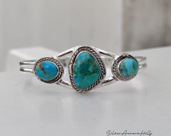 Pode incluir: Pulseira de punho de prata com três pedras turquesas. A pedra central tem uma forma irregular, ladeada por duas pedras redondas. A pulseira tem um acabamento polido e um design aberto. Medidas em centímetros.