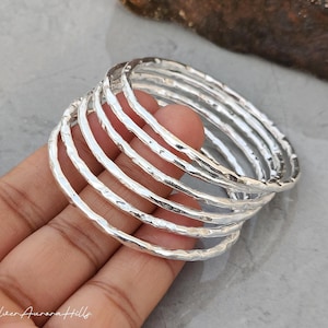 May include: A stack of silver bangles with a hammered texture. The bracelets are round and shiny, reflecting light. The image is taken from a close-up angle, showing the details of the metalwork. The bangles are stacked on top of each other.