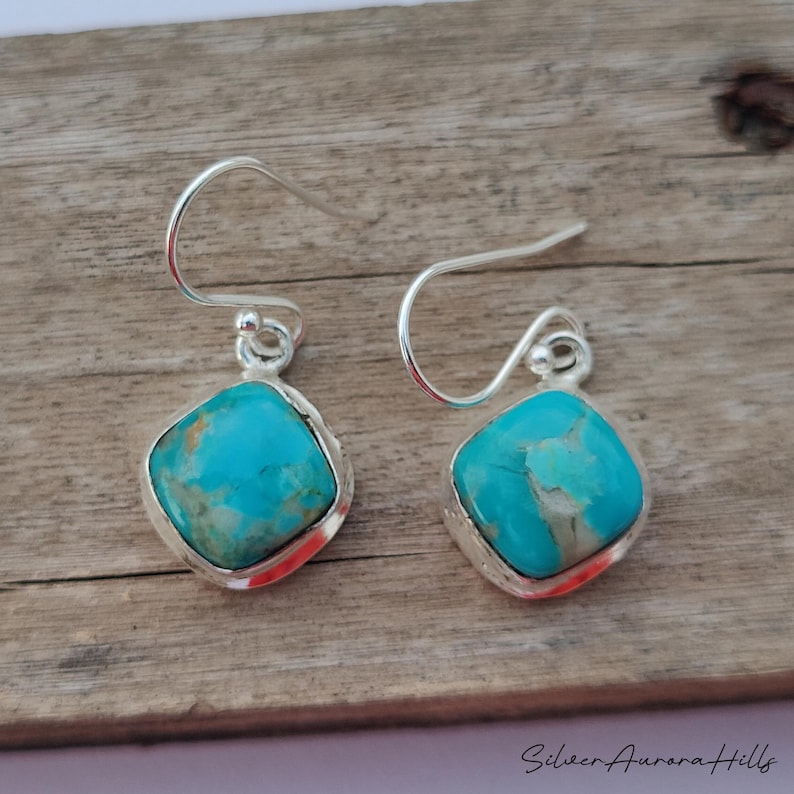 May include: A pair of silver drop earrings with square turquoise stones. The turquoise stones have a marbled appearance with shades of blue and green. The earrings are set in a silver bezel and have a simple hook design.
