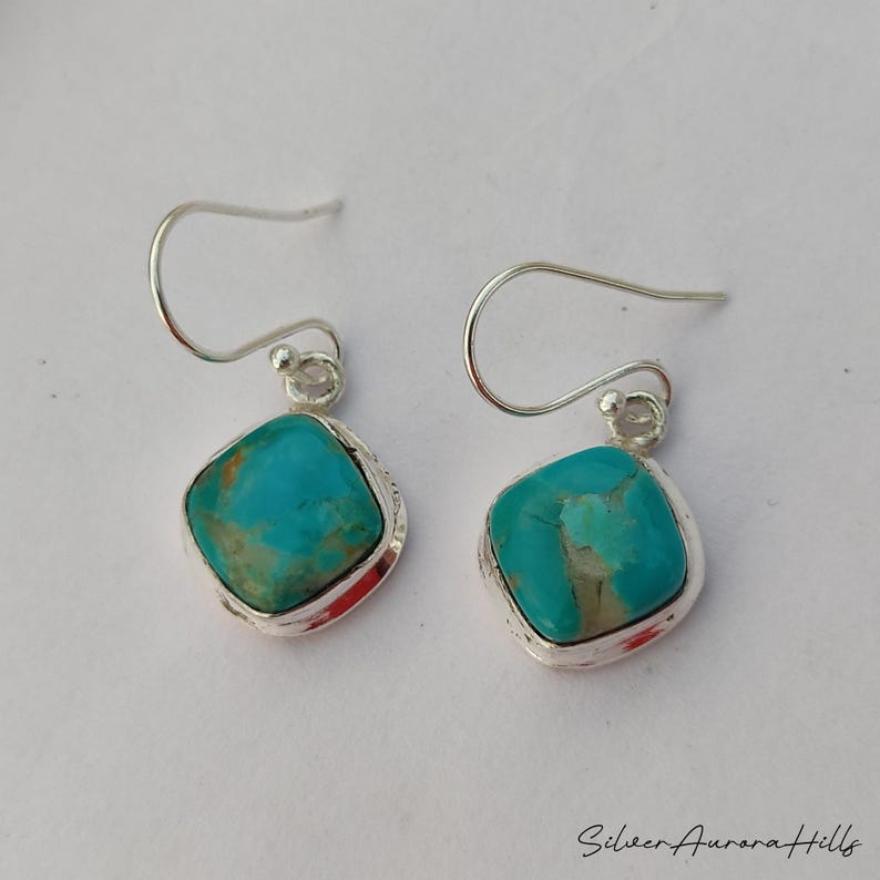 May include: A pair of turquoise earrings with a square, rounded shape. The turquoise stones are set in a silver bezel with a simple hook closure. The earrings are displayed on a white background.