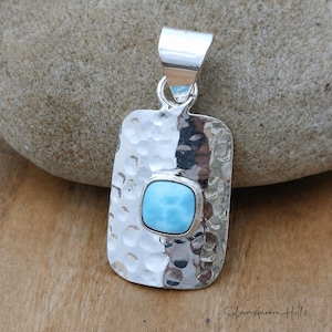 May include: A rectangular silver pendant with a hammered texture and a light blue square gemstone. The pendant has a polished bail for a chain. The gemstone is set in a bezel setting. The pendant is approximately 1.25 inches long.
