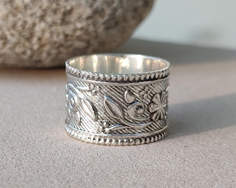 May include: A wide, silver-colored ring with an intricate floral design. The ring features embossed leaves, flowers, and swirling patterns. The band has a textured surface and a beaded border. The ring is a statement piece of jewelry.