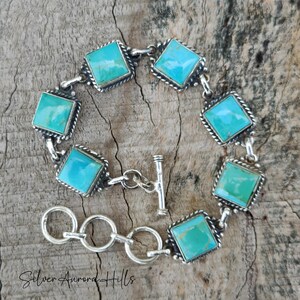 May include: A silver bracelet featuring square turquoise stones set in a decorative frame. The bracelet has a toggle clasp and is displayed on a wooden surface. The text "Silver Aurora Hills" is visible at the bottom.