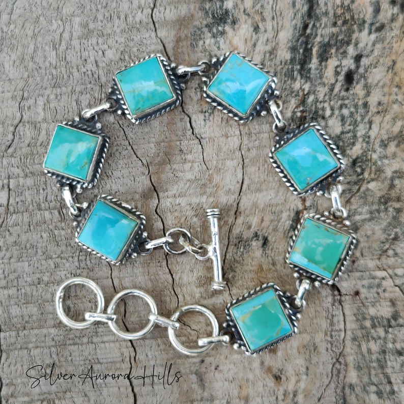 May include: A silver bracelet featuring square turquoise stones set in a decorative frame. The bracelet has a toggle clasp and is displayed on a wooden surface. The text "Silver Aurora Hills" is visible at the bottom.