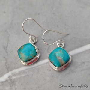 May include: A pair of turquoise drop earrings. Each earring features a square-shaped, vibrant blue-green stone set in a silver bezel. The earrings have a simple hook design for pierced ears. The earrings are displayed on a gray surface.
