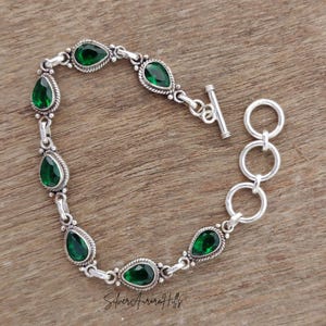 May include: A silver bracelet with pear-shaped, emerald-green gemstones. The bracelet features a toggle clasp and is set against a wooden background. The gemstones are framed by a decorative silver border.