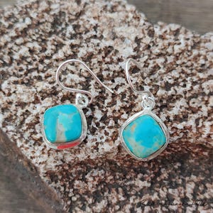 May include: A pair of turquoise and silver earrings. Each earring features a square, light blue stone with brown veining, set in a silver bezel. The earrings have a simple hook design. The background is a textured, brown surface.