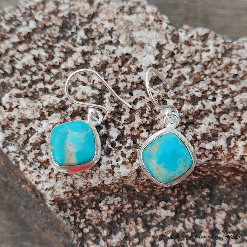 May include: A pair of turquoise and silver earrings. Each earring features a square, light blue stone with brown veining, set in a silver bezel. The earrings have a simple hook design. The background is a textured, brown surface.