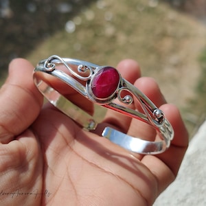 May include: A silver bangle bracelet with a large, oval, deep red gemstone. The bracelet has decorative swirls and small ball accents. The open design allows for easy wear. The bracelet is shown on a hand.