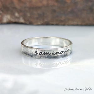 Peut inclure: Une bague anneau en argent avec une finition polie et une section inférieure texturée et assombrie. La phrase "i am enough" est gravée en écriture cursive noire sur la surface extérieure de l'anneau, créant un contraste net avec le métal.