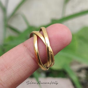 May include: A gold-colored ring with three intertwined bands. The ring is shown on a finger, with a blurred green background. The ring has a smooth, polished finish and a simple, elegant design.