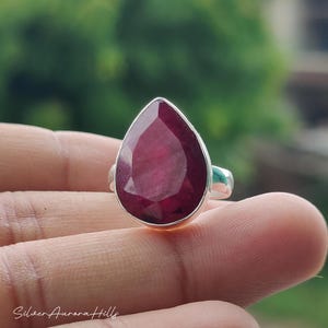 May include: A silver ring with a large, teardrop-shaped, deep red gemstone. The gemstone is cut to reflect light. The ring has a plain silver band. The background is a blurred green, possibly an outdoor scene.