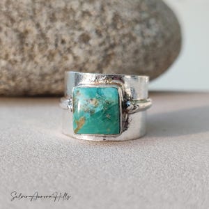 May include: A wide silver ring featuring a square turquoise stone. The turquoise stone displays a mottled pattern of green and brown. The ring has a textured band and a polished finish. The words "Silver Aurora Hills" are at the bottom.