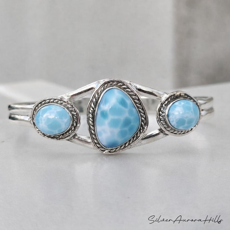 Op de afbeelding: Zilveren armband met drie lichtblauwe larimar stenen. De centrale steen heeft een onregelmatige vorm, geflankeerd door twee ovale stenen. De armband heeft een delicaat, open ontwerp met een gedraaide touwrand rond de stenen.