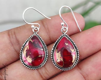 May include: Silver drop earrings with a teardrop-shaped red and gold stone. The stone is framed by a twisted silver border. The earrings have a simple hook closure.