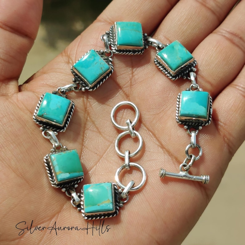 May include: A silver bracelet featuring square turquoise stones. Each stone is set in a square frame with a twisted rope design. The bracelet has a toggle clasp and is displayed on a hand.