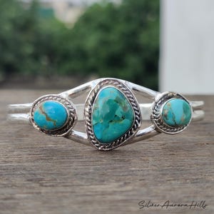 Op de afbeelding: Een zilveren manchetarmband met drie turkooizen stenen. De centrale steen heeft een onregelmatige vorm, geflankeerd door twee ronde stenen. De turkoois heeft een gevlekte blauwgroene kleur. De armband is geplaatst tegen een vage natuurlijke achtergrond.