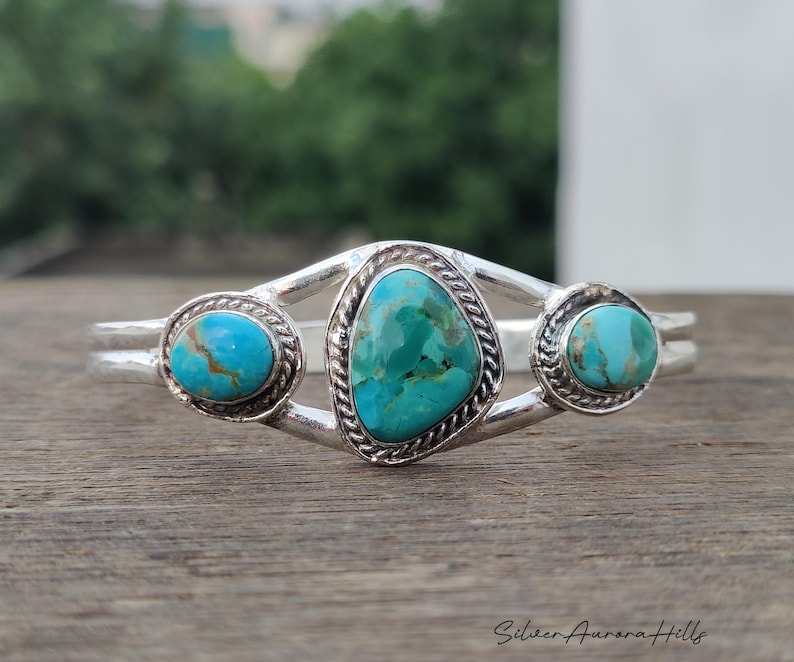 Op de afbeelding: Een zilveren manchetarmband met drie turkooizen stenen. De centrale steen heeft een onregelmatige vorm, geflankeerd door twee ronde stenen. De turkoois heeft een gevlekte blauwgroene kleur. De armband is geplaatst tegen een vage natuurlijke achtergrond.