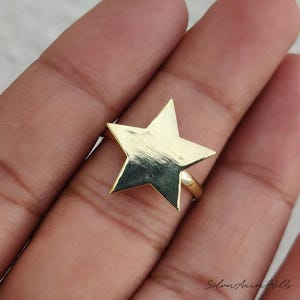 May include: A gold-toned ring featuring a large, flat, five-pointed star as the centerpiece. The star has a smooth, reflective surface with subtle horizontal texture lines. The band is thin and circular, connecting to the back of the star.
