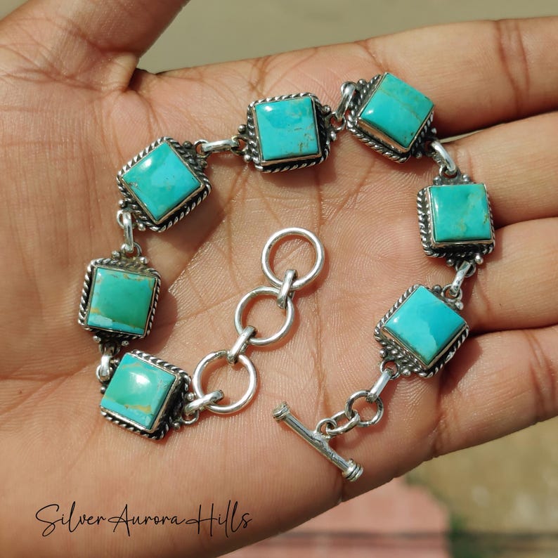 May include: A silver bracelet featuring square turquoise stones. Each stone is set in a decorative silver frame with a twisted rope design. The bracelet has a toggle clasp.