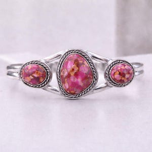 Op de afbeelding: Zilverkleurige manchetarmband met drie roze edelstenen. De edelstenen hebben een gespikkelde uitstraling met tinten roze, rood en bruin. De armband heeft een gevlochten touwontwerp rond elke steen.