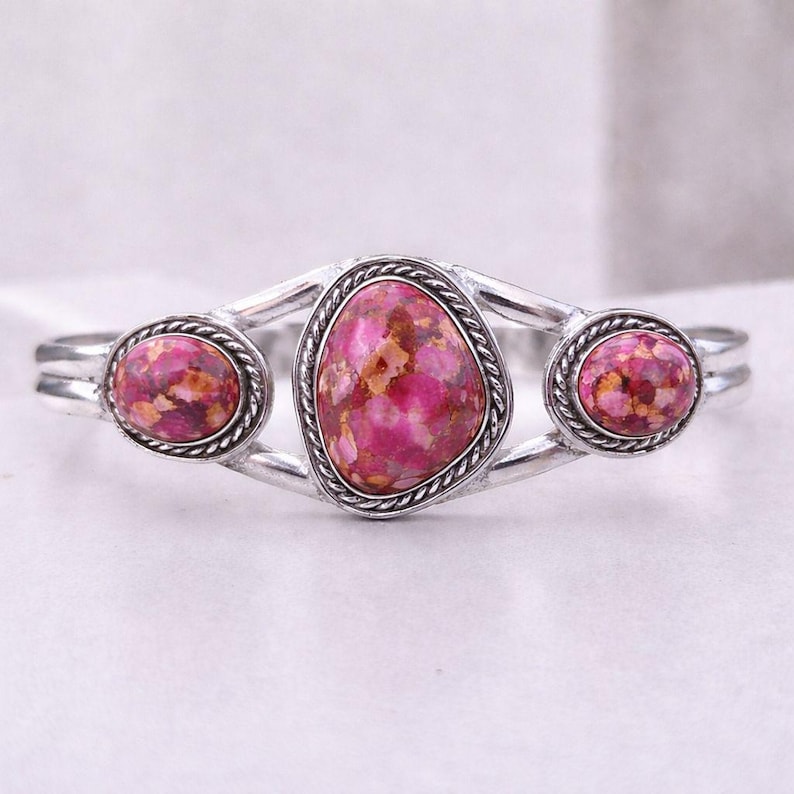 Op de afbeelding: Zilverkleurige manchetarmband met drie roze edelstenen. De edelstenen hebben een gespikkelde uitstraling met tinten roze, rood en bruin. De armband heeft een gevlochten touwontwerp rond elke steen.