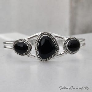 Op de afbeelding: Een zilveren armband met drie zwarte onyx stenen. De centrale steen is driehoekig, geflankeerd door twee ovale stenen. De armband heeft een gedetailleerd, getextureerd ontwerp rond de stenen.