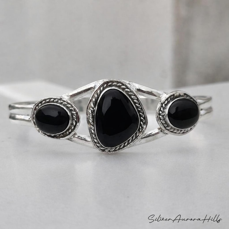 Op de afbeelding: Een zilveren armband met drie zwarte onyx stenen. De centrale steen is driehoekig, geflankeerd door twee ovale stenen. De armband heeft een gedetailleerd, getextureerd ontwerp rond de stenen.