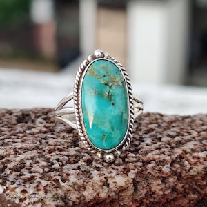 May include: Oval turquoise ring with a silver band. The turquoise stone is a vibrant teal color with brown veining. The ring has a twisted rope design around the stone and a split band. The ring is set on a textured brown surface.
