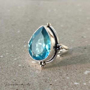 May include: A silver ring featuring a large, pear-shaped, light blue gemstone. The ring has a detailed, ornate setting with decorative elements on the sides. The gemstone is faceted, reflecting light. The ring is displayed on a neutral surface.
