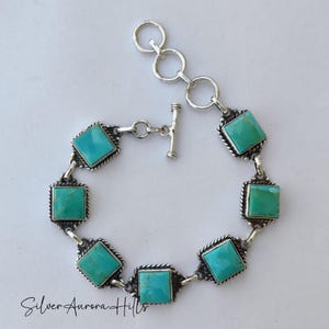 May include: A silver bracelet featuring square turquoise stones. The bracelet has a toggle clasp and is connected by silver links. Each turquoise stone is set in a square frame with a twisted rope design. The text "Silver Aurora Hills" is at the bottom.