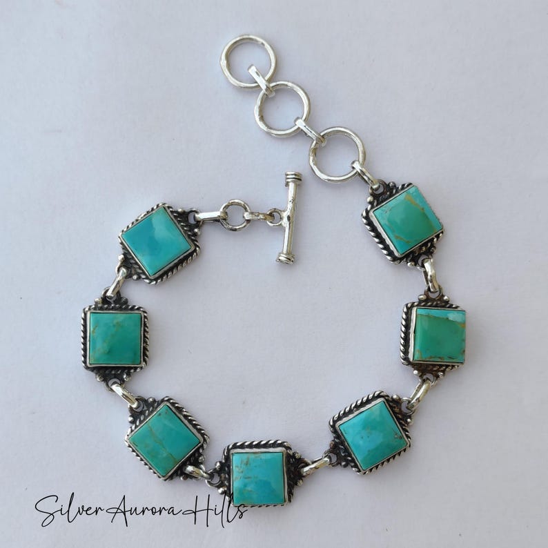 May include: A silver bracelet featuring square turquoise stones. The bracelet has a toggle clasp and is connected by silver links. Each turquoise stone is set in a square frame with a twisted rope design. The text "Silver Aurora Hills" is at the bottom.