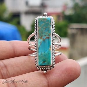 May include: A rectangular turquoise ring with a silver band. The turquoise stone is a vibrant blue-green color with brown veining. The ring has a braided border and three bands on each side.